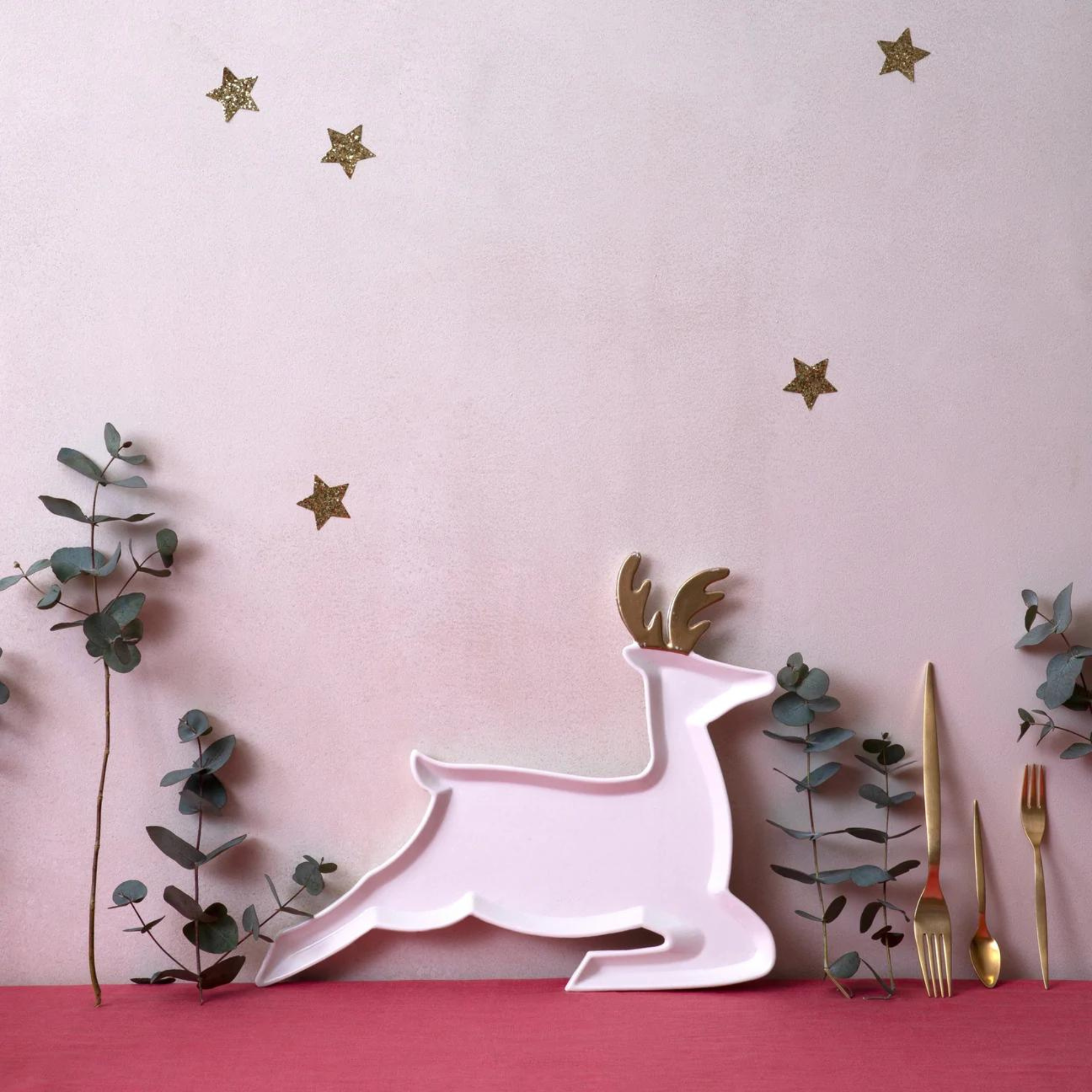 White reindeer platter next to green eucalyptus leaves and golden cutlery on a pink background with gold stars.