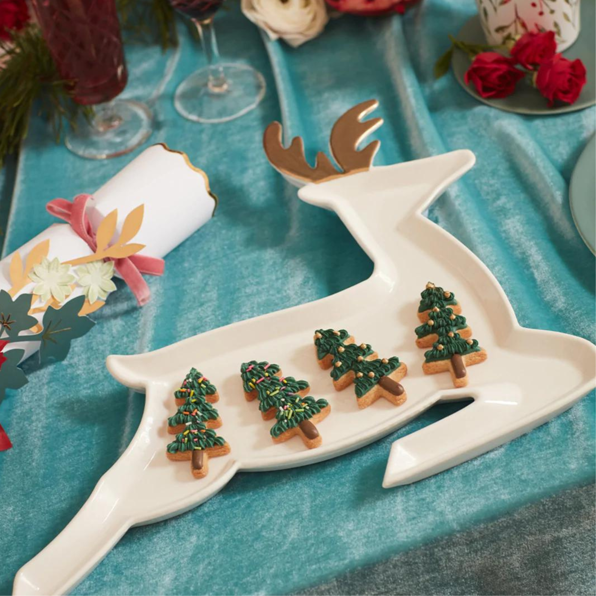White reindeer platter with decorated tree-shaped cookies, set on a teal tablecloth with festive decorations.