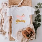 White baby onesie with "Sweet as Honey" text, surrounded by greenery, wooden beads, and soft fabric.