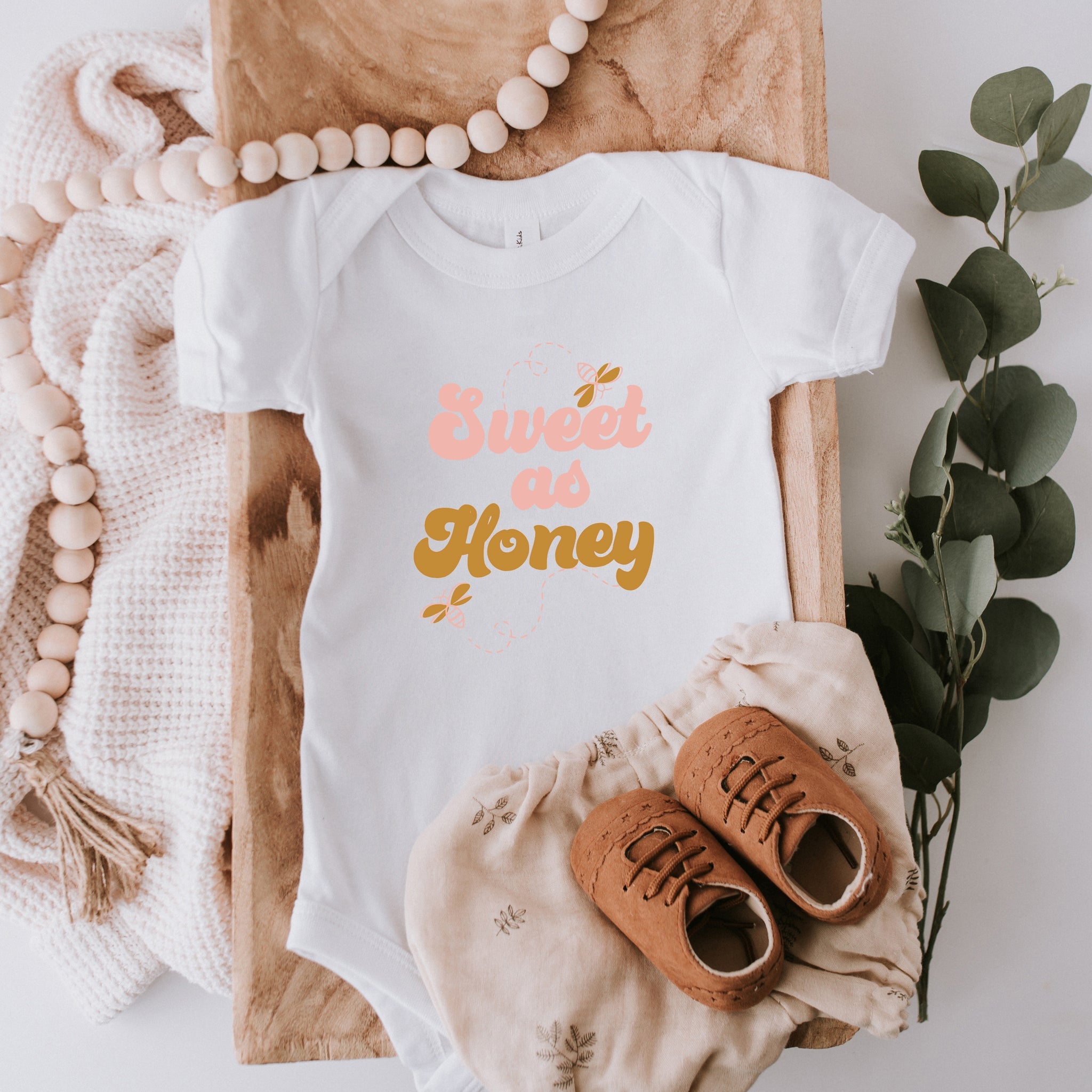 White baby onesie with "Sweet as Honey" text, surrounded by greenery, wooden beads, and soft fabric.