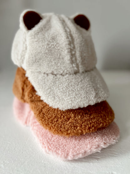 Fuzzy bear-themed cap stacked on soft, colorful fleece hats in shades of brown and pink against a neutral background.