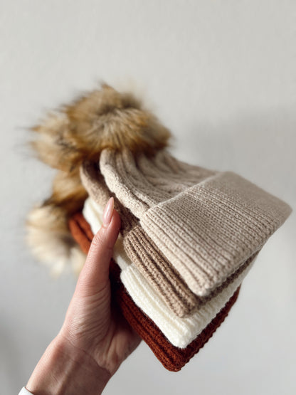 Stack of knitted beanies in beige, brown, cream, and rust colors, held in a hand against a neutral background.