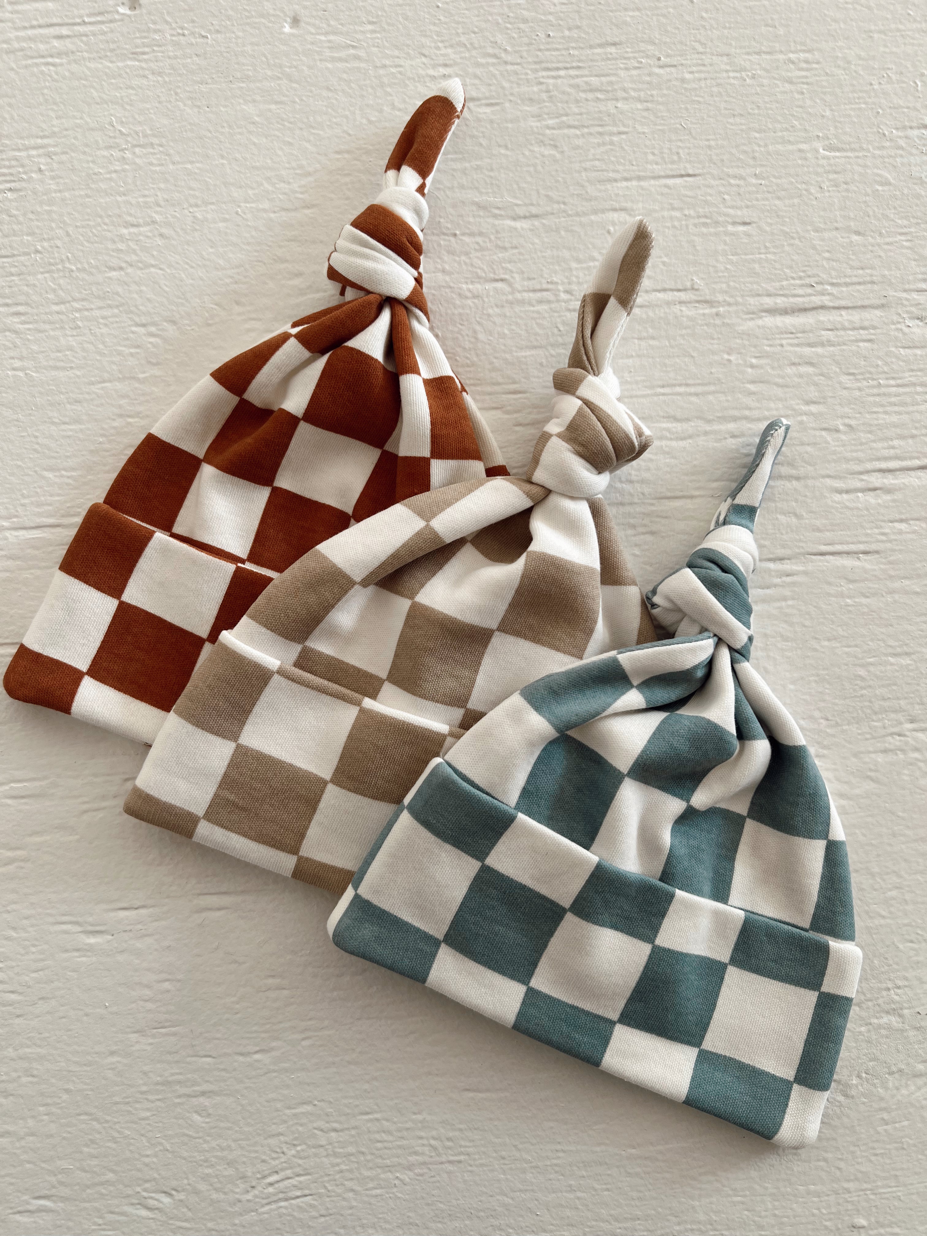 Three checkered beanies in rust, beige, and teal colors displayed against a plain white background.