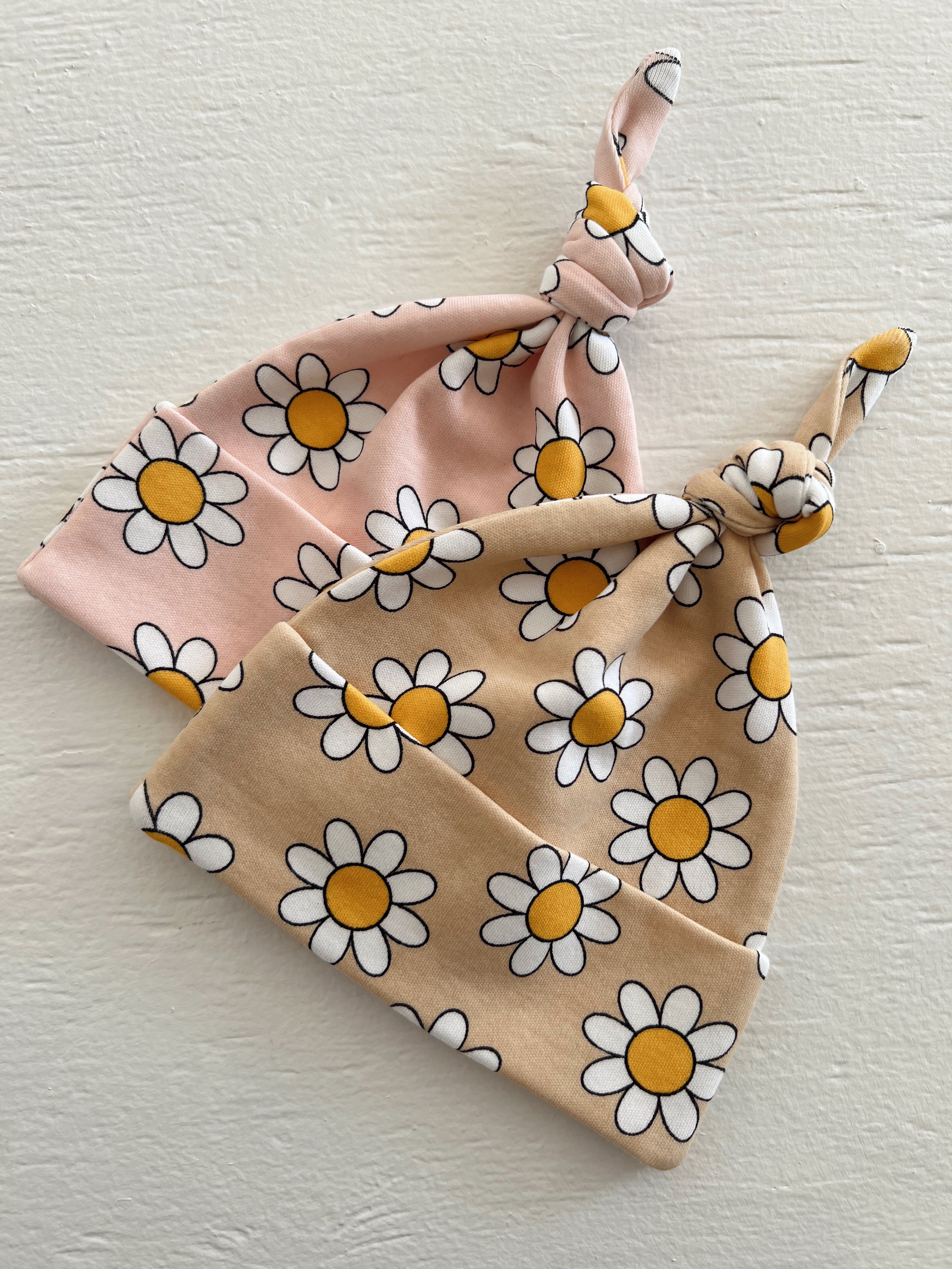 Two floral baby hats in pink and tan with yellow daisies, stacked on a light textured surface.