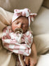 Newborn baby sleeping in a pink checkered outfit with a matching headband and pacifier, held in a caregiver's arms.