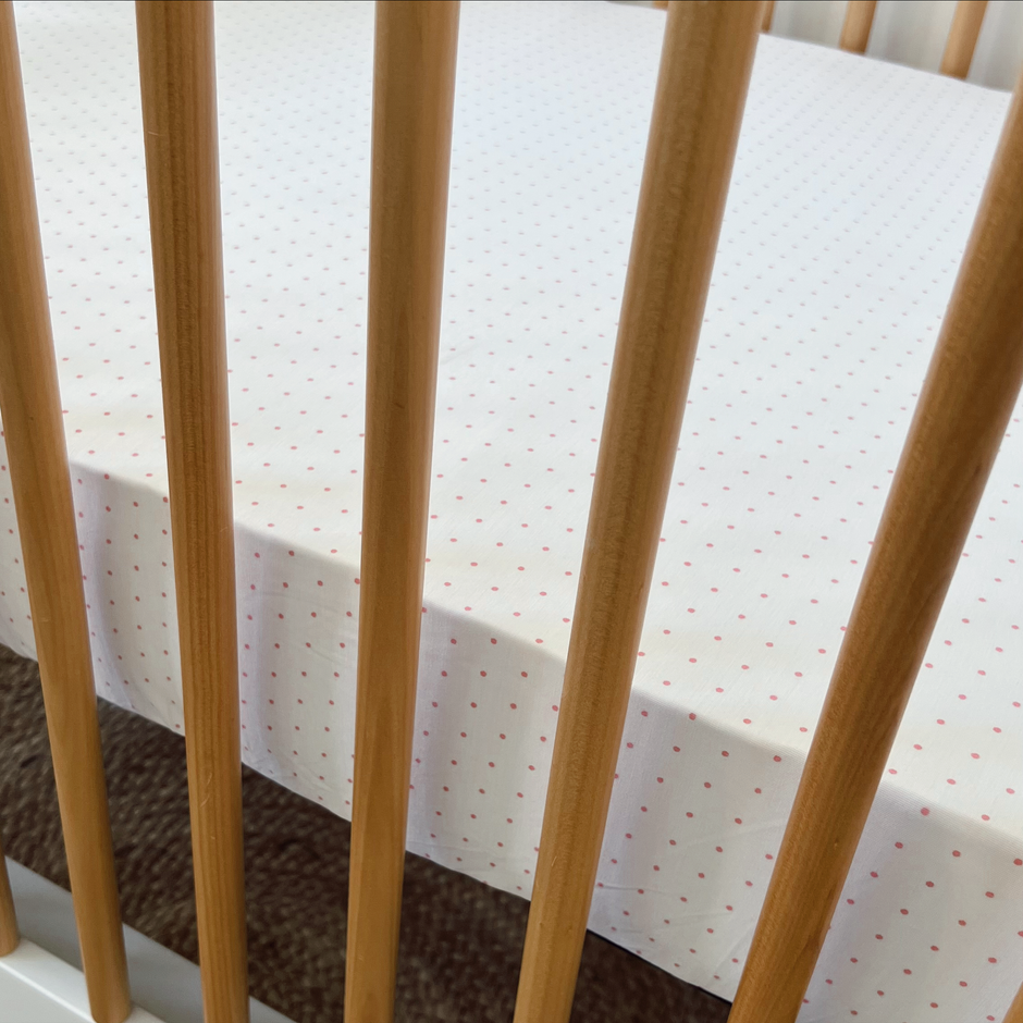 Crib with soft white bedding featuring small pink polka dots, seen through wooden slats.