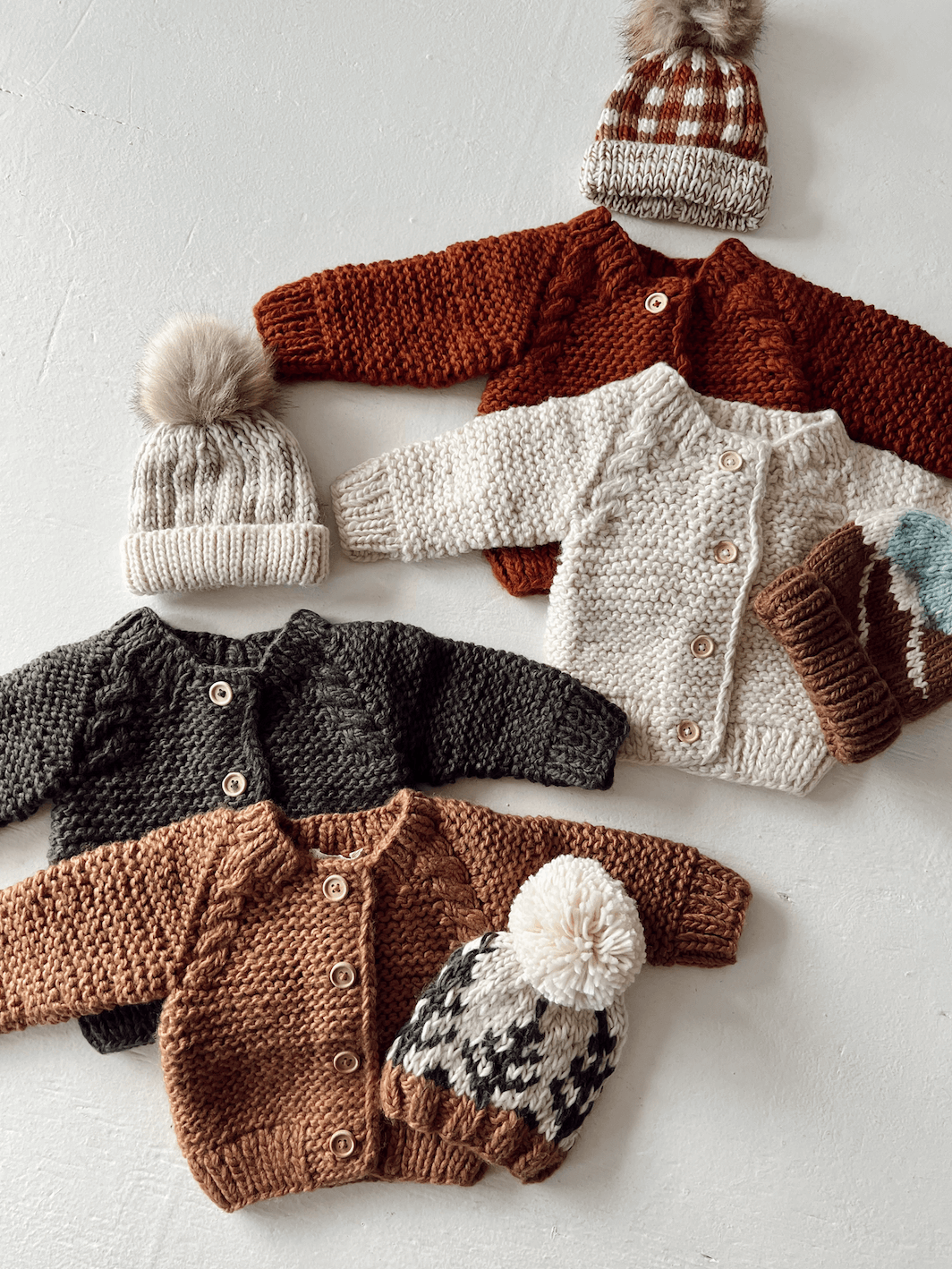 Knitted baby sweaters and hats in various colors, featuring buttons and playful pom-poms on top.