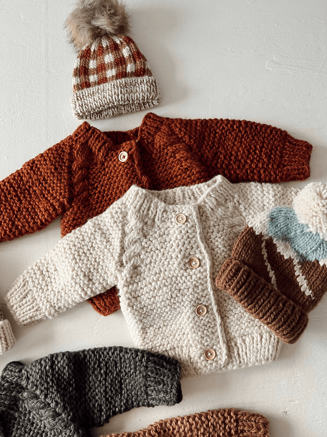 Knitted baby sweaters in various colors with a checked hat, arranged on a light surface.