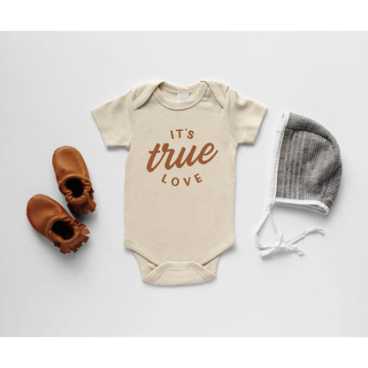Beige baby onesie with "It's true love" text, paired with brown baby shoes and a gray bonnet, against a white background.